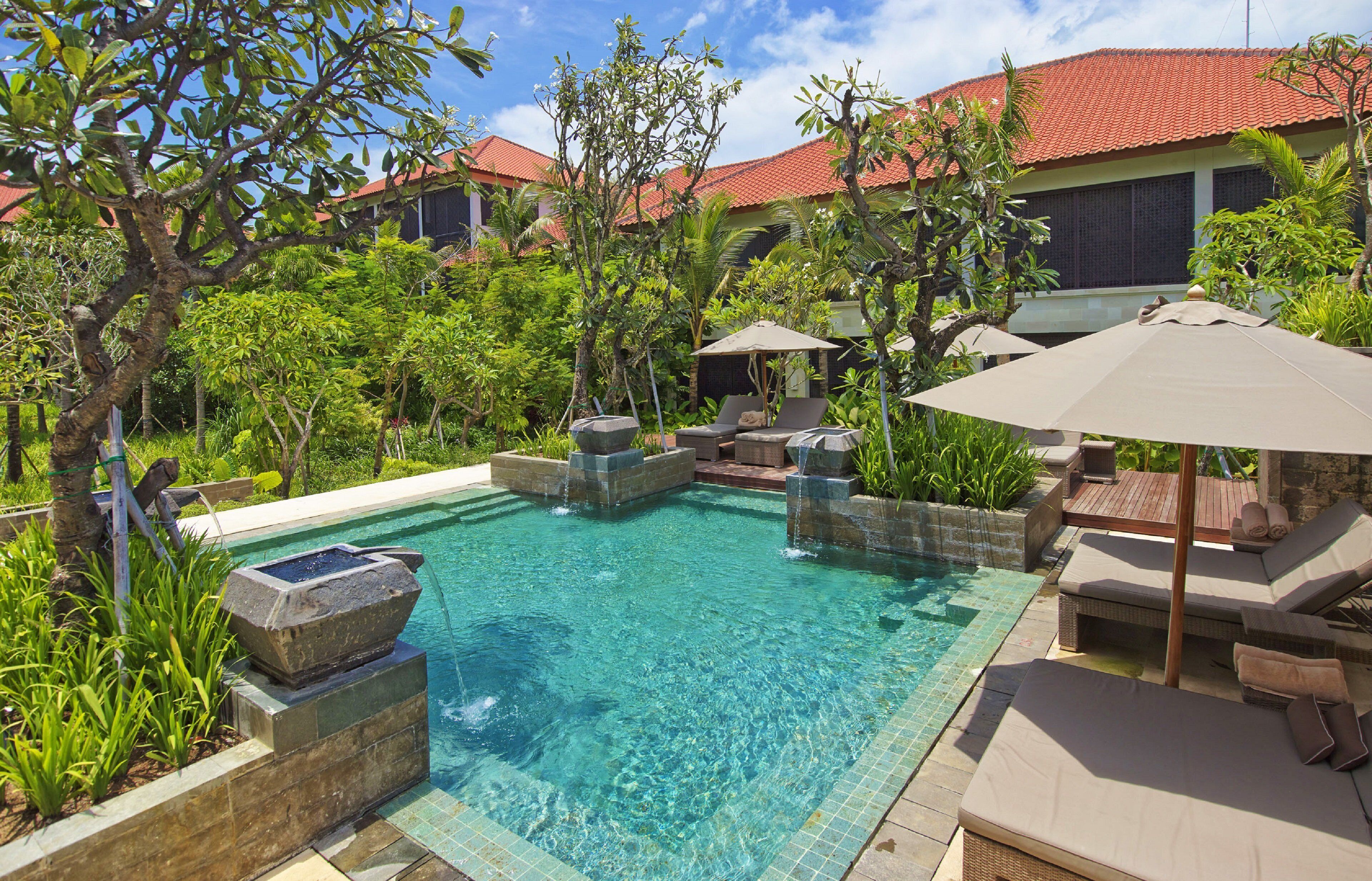 A pool with clear water, surrounded by lounge chairs and umbrellas, with a red-roofed building in the background.