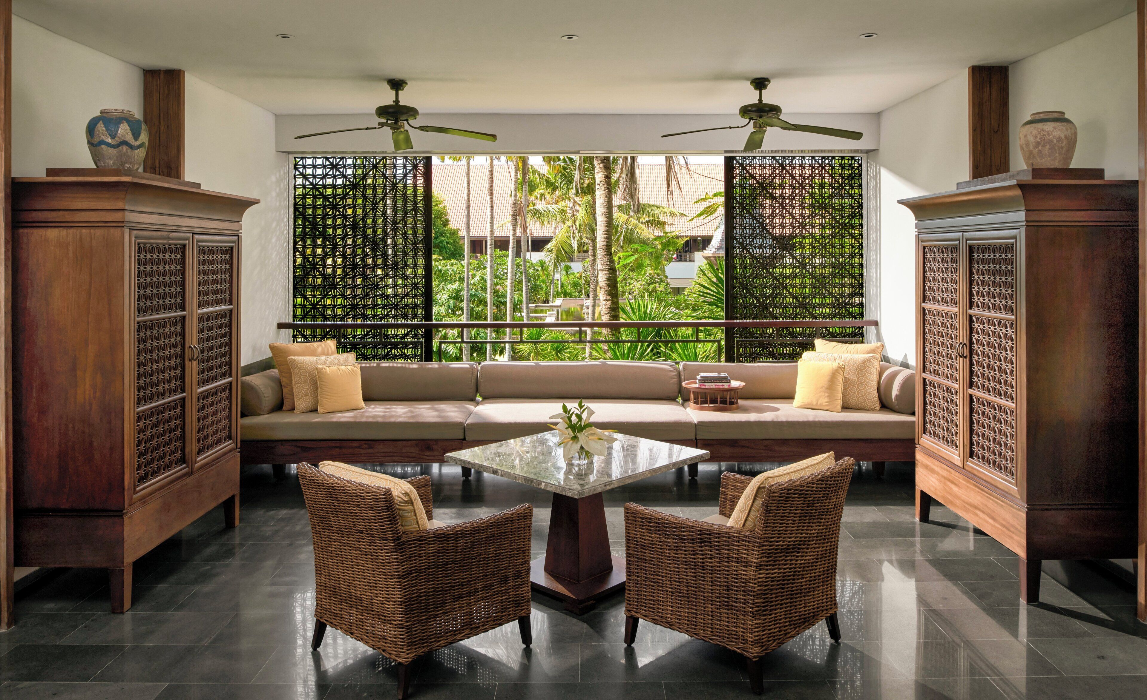 A room with a sofa, two wicker chairs, a glass table, ceiling fans, and a view through a patterned screen.