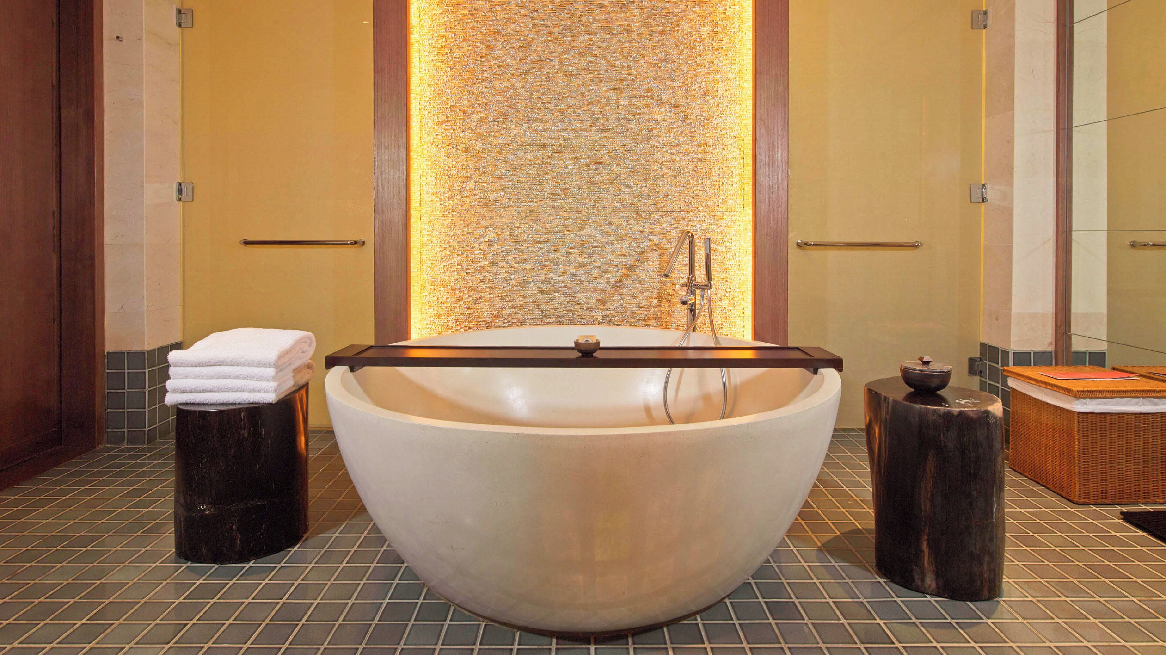 A large freestanding bathtub in a bathroom with a textured wall behind it, a towel rack, and a small wooden stool.