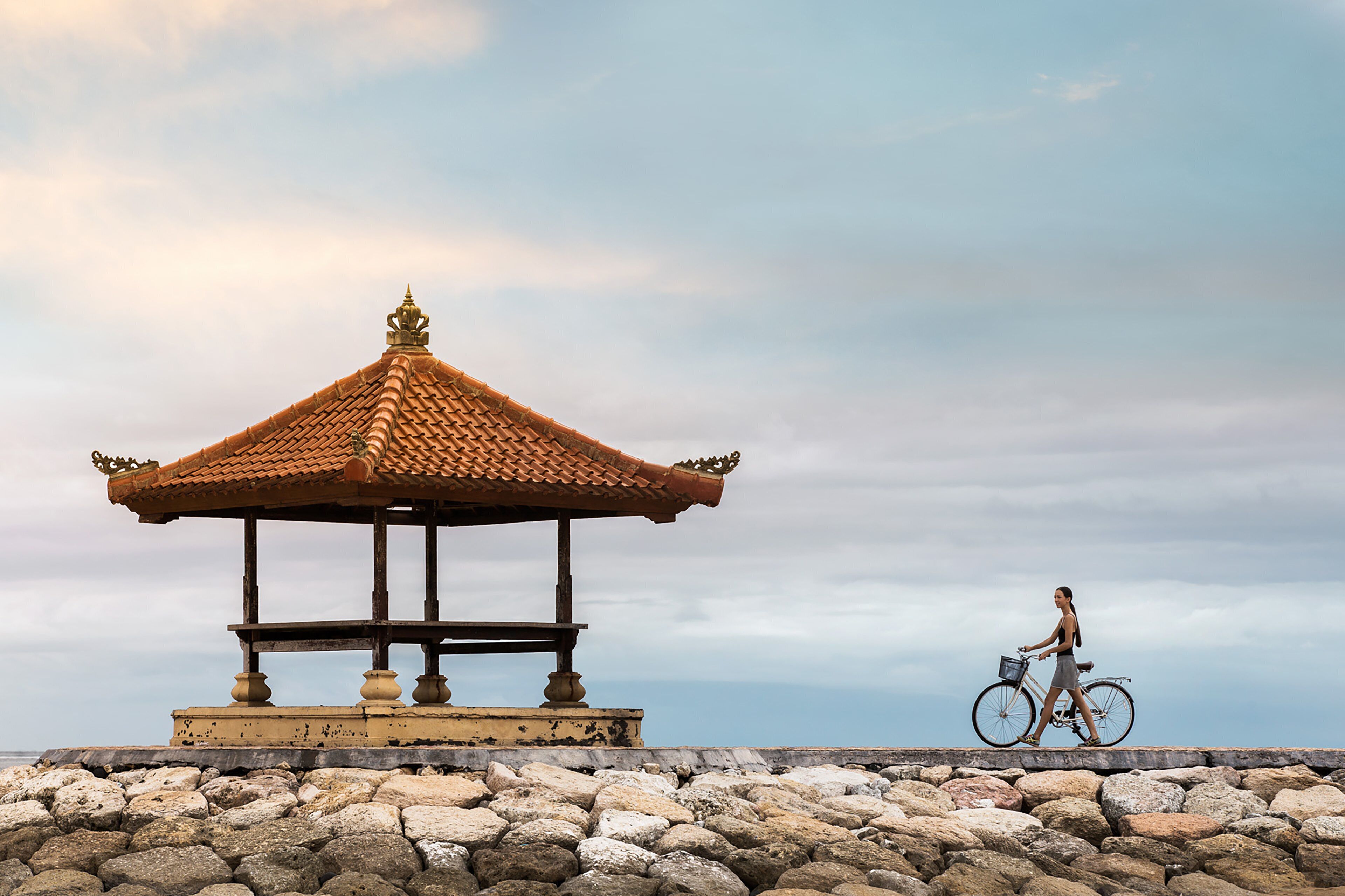 A gazebo with a tiled roof and a stone base, located on a rocky platform with a view of the sea.