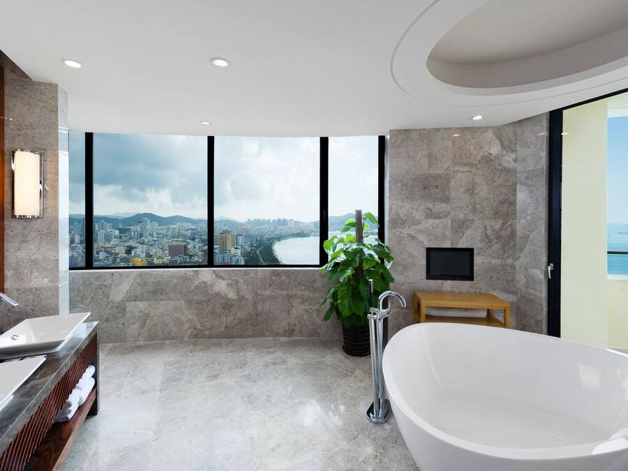 A modern bathroom with a large bathtub, a vanity with two sinks, a potted plant, and a city view through large windows.