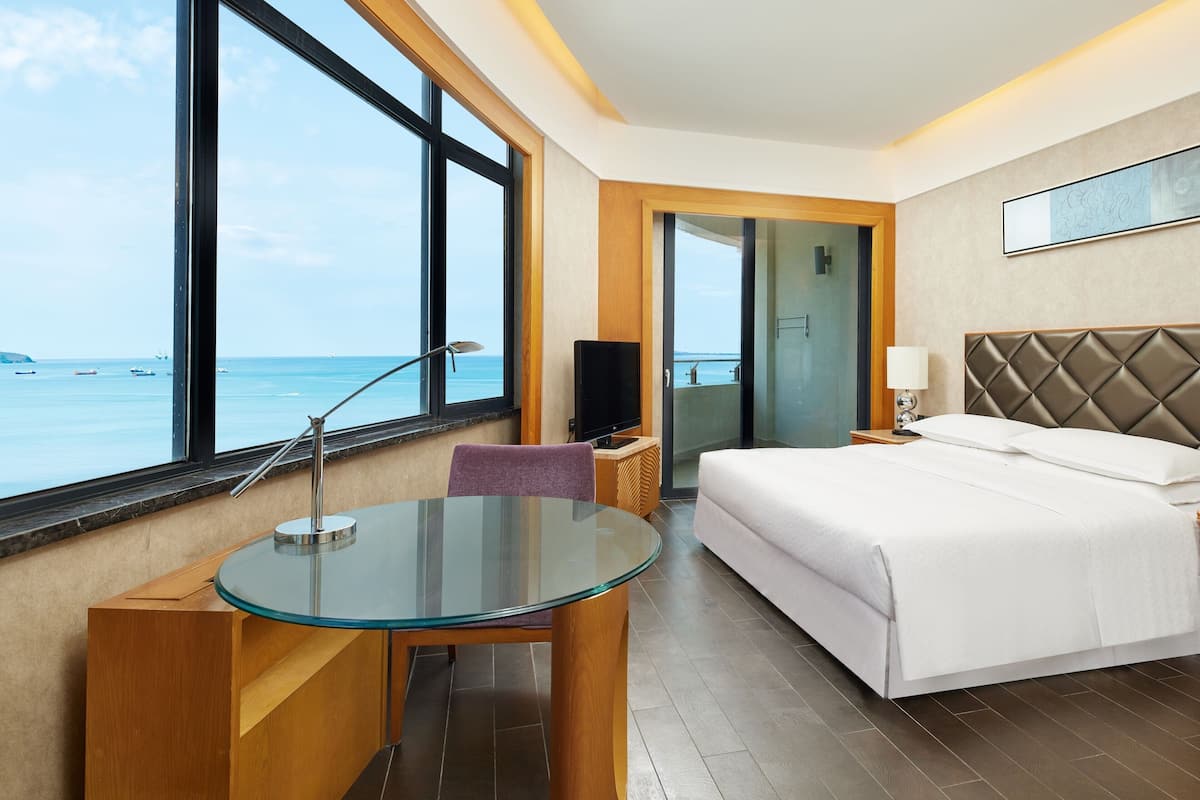 A hotel room with a bed, a glass table, a TV, and a view of the sea.