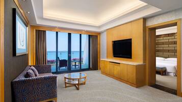 A modern hotel room with a sofa, a TV, a small table, and a balcony with a view of the sea.
