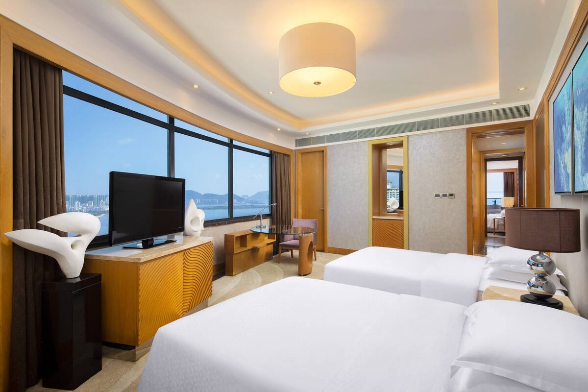 A hotel room with two beds, a TV, a desk, a chair, and a large window offering a city view.