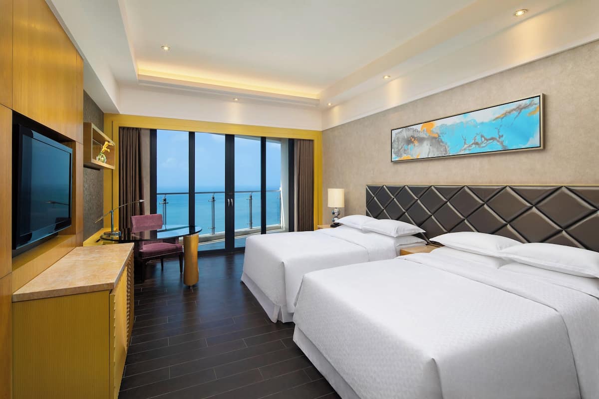 A hotel room with two beds, a TV, a desk, a chair, and a large window with a view of the sea.