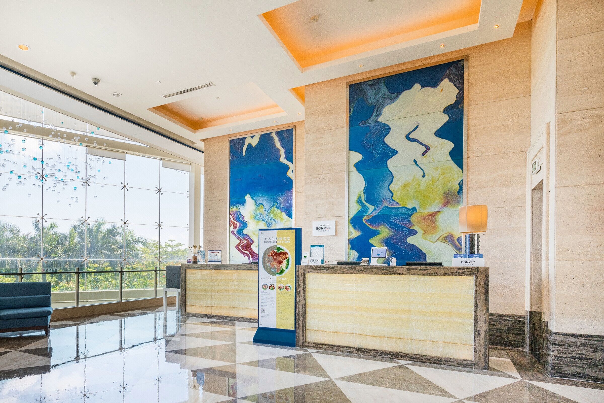 Um saguão de hotel com balcão de recepção, uma janela grande com vista para área verde e um design moderno no teto.