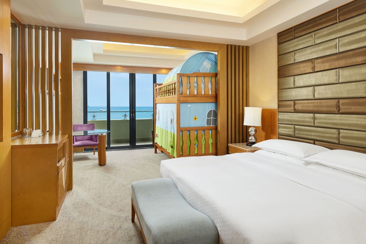 A hotel room with a large bed, a wooden headboard, a balcony with a view of the sea, a desk with a chair, and a small table with a lamp.