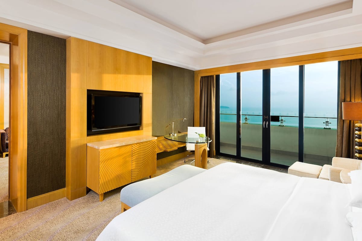 A hotel room with a large bed, a TV, a desk with a glass top, and a view of the sea.