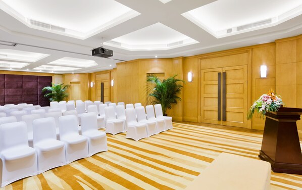 A conference room with rows of white chairs, a podium with a floral arrangement, and wooden paneling on the walls.