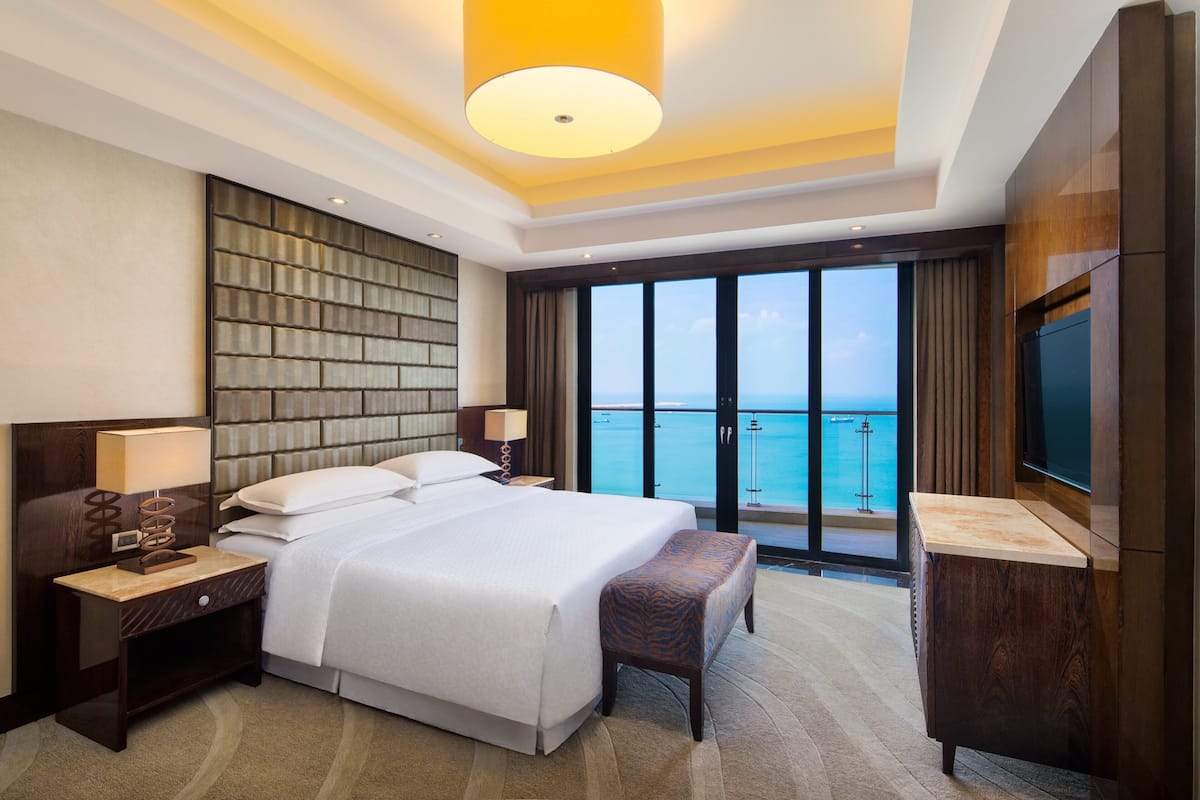 A hotel room with a large bed, a desk with a lamp, a television, and a view of the ocean.