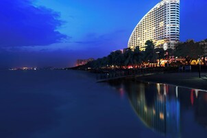 Un hotel moderno y grande con fachada curva, situado cerca de un frente marítimo con una masa de agua tranquila que refleja sus luces.