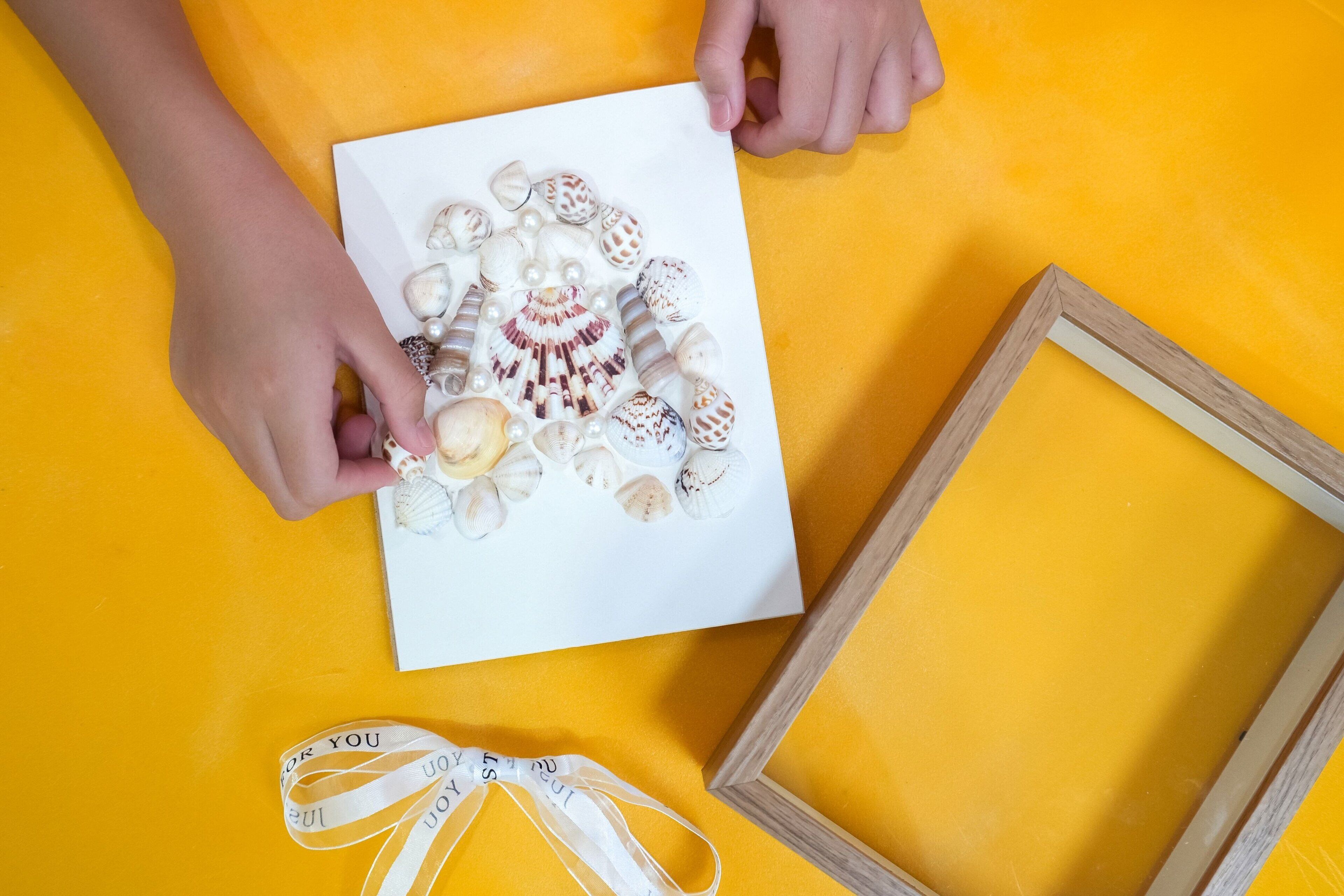 Una mano sosteniendo una tarjeta blanca con caracolas y perlas, un marco de madera y una cinta con la palabra 'YOU'.
