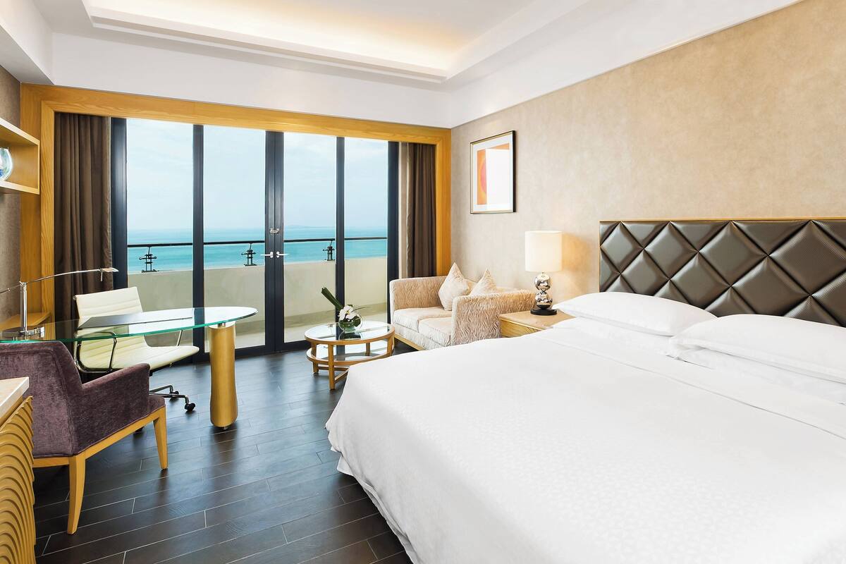 A hotel room with a large bed, a desk with a chair, a glass table, and a view of the sea through large windows.