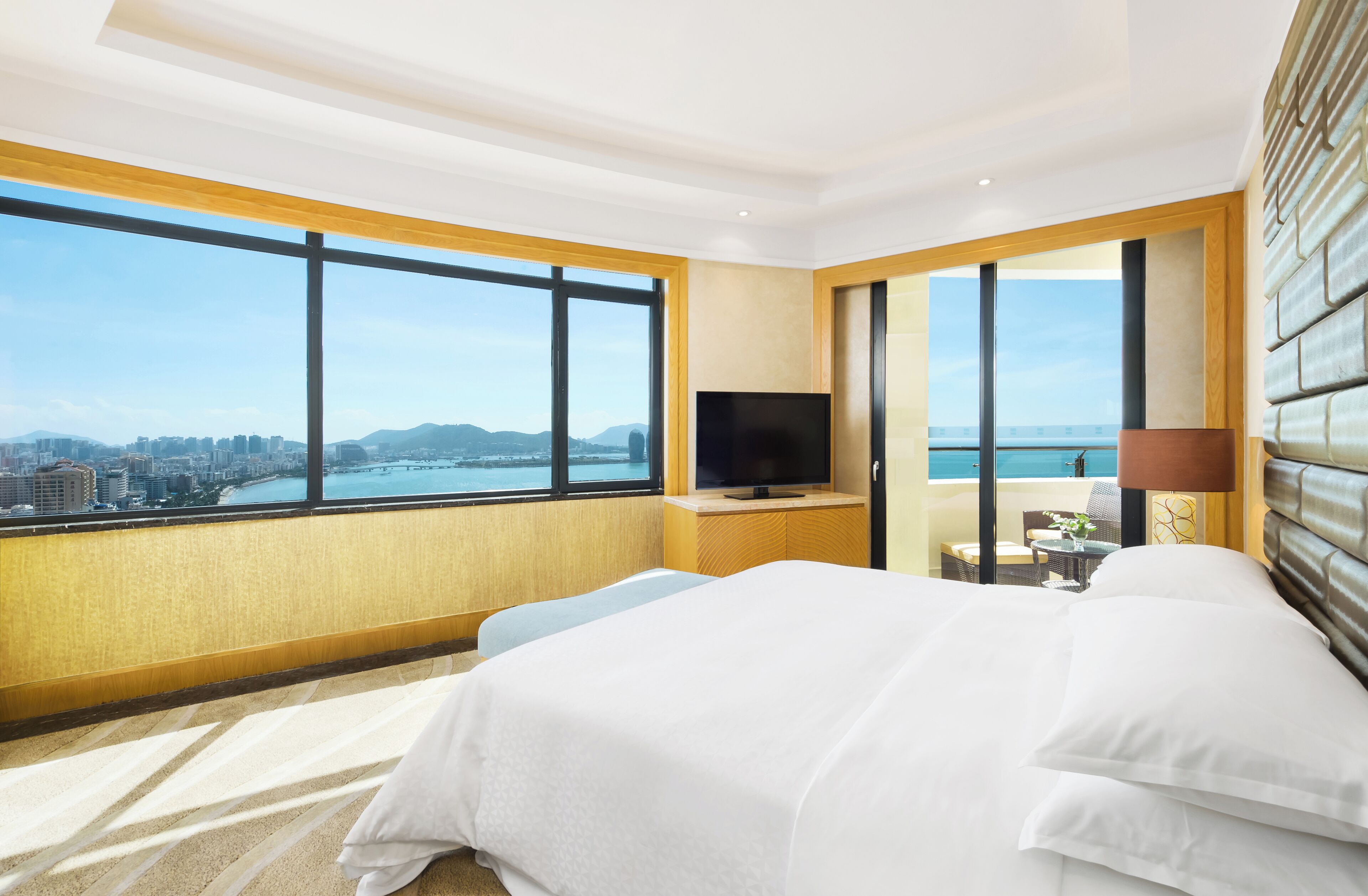 A hotel room with a large window offering a city and ocean view, a bed, a TV, and a small seating area.