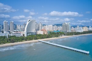 A coastal cityscape with high-rise buildings, a beach, and a pier extending into the water.