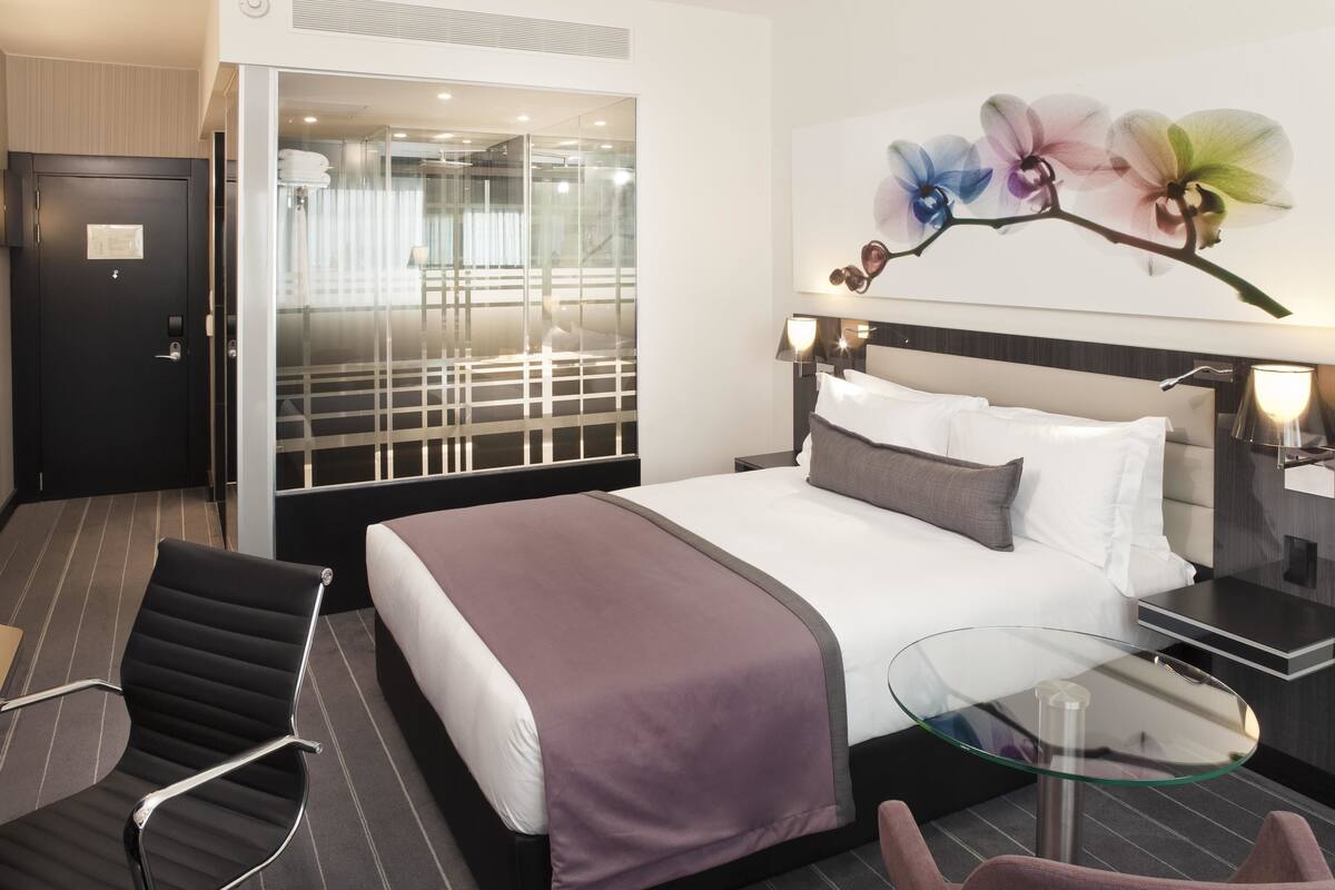 Une chambre d’hôtel moderne avec un grand lit, une table en verre, une chaise et une vue sur l’intérieur de l’hôtel grâce à une paroi vitrée.