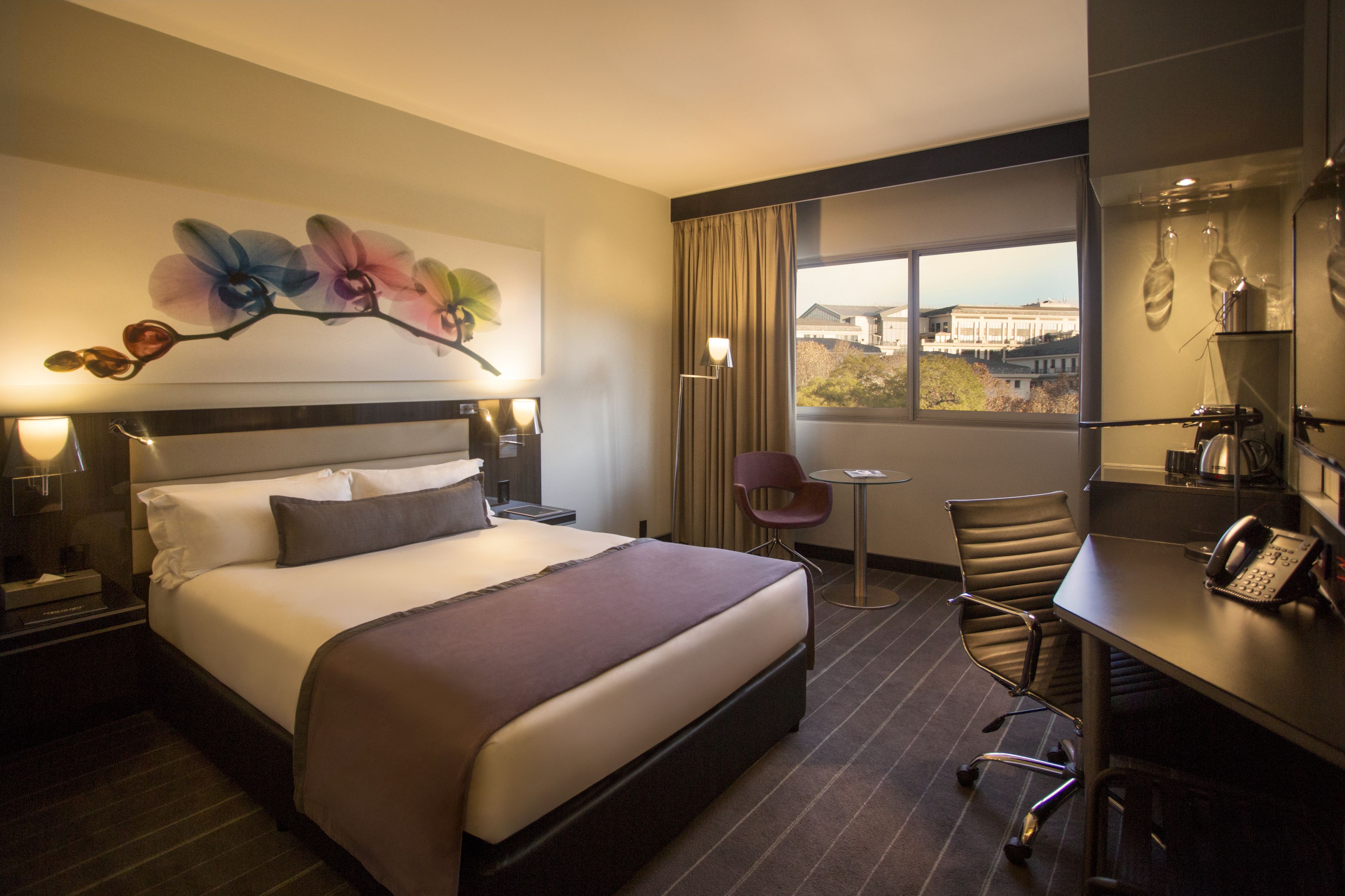 A hotel room with a large bed, a desk, a chair, a small table, and a window with a view of buildings.