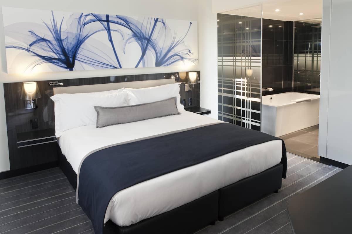 Une chambre moderne avec un grand lit, des lampes de chevet, une œuvre d’art abstraite accrochée au mur, et une salle de bain séparée dotée d’une baignoire.