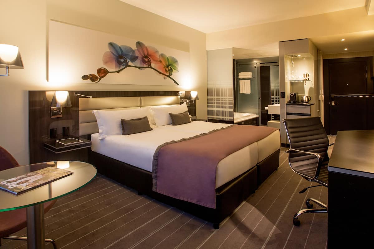 Une chambre d’hôtel avec un grand lit, un bureau, une chaise, une table en verre et un tableau au mur.