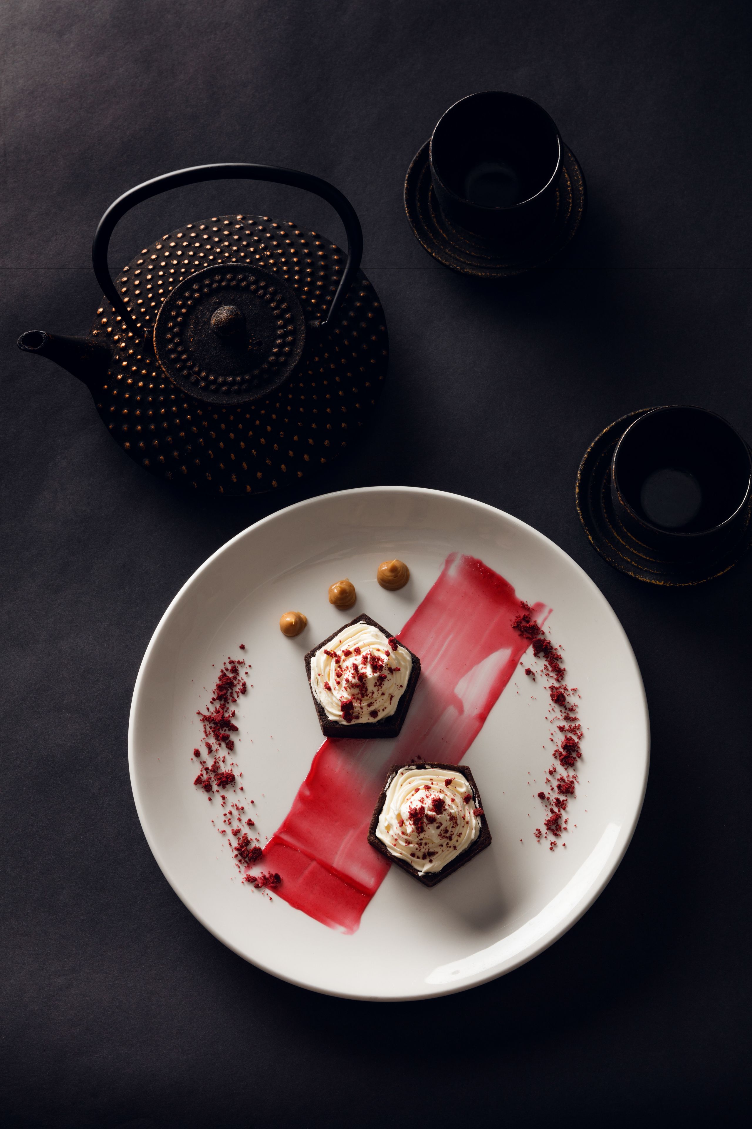 A dark surface with a white plate containing a dessert, a teapot, and two cups.