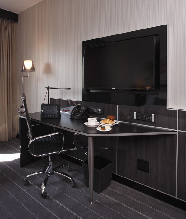 A modern hotel room with a large flat-screen TV, a desk with a phone, a lamp, and a small breakfast setup.