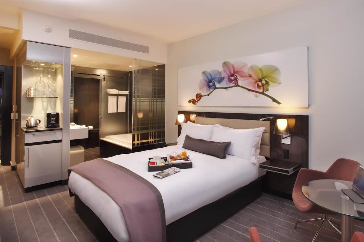 Une chambre d’hôtel moderne dotée d’un grand lit, d’une table de chevet et d’une cloison en verre qui sépare la chambre de la salle de bain.