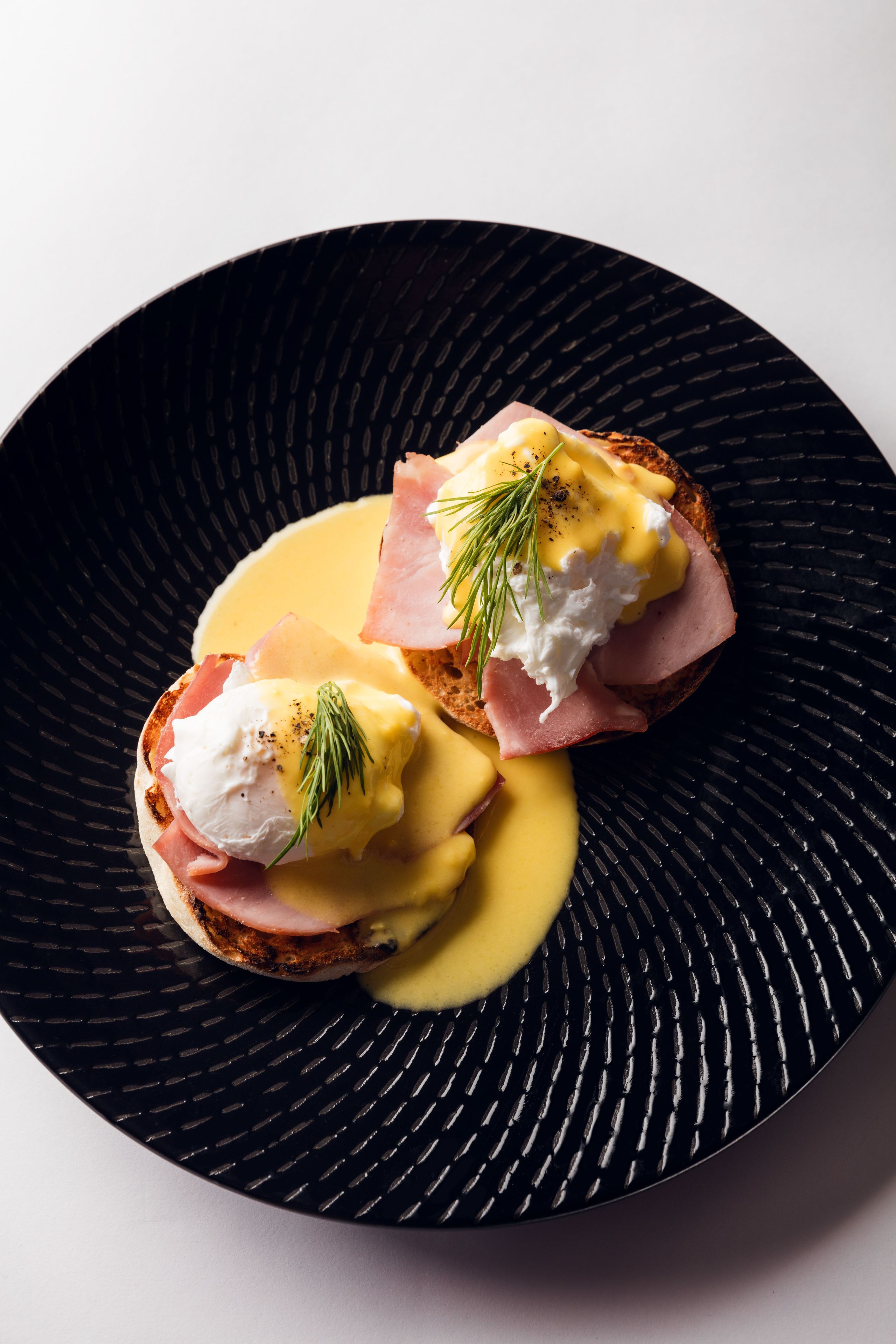 A black plate with two pieces of toast topped with ham, poached eggs, and hollandaise sauce, garnished with a sprig of dill.