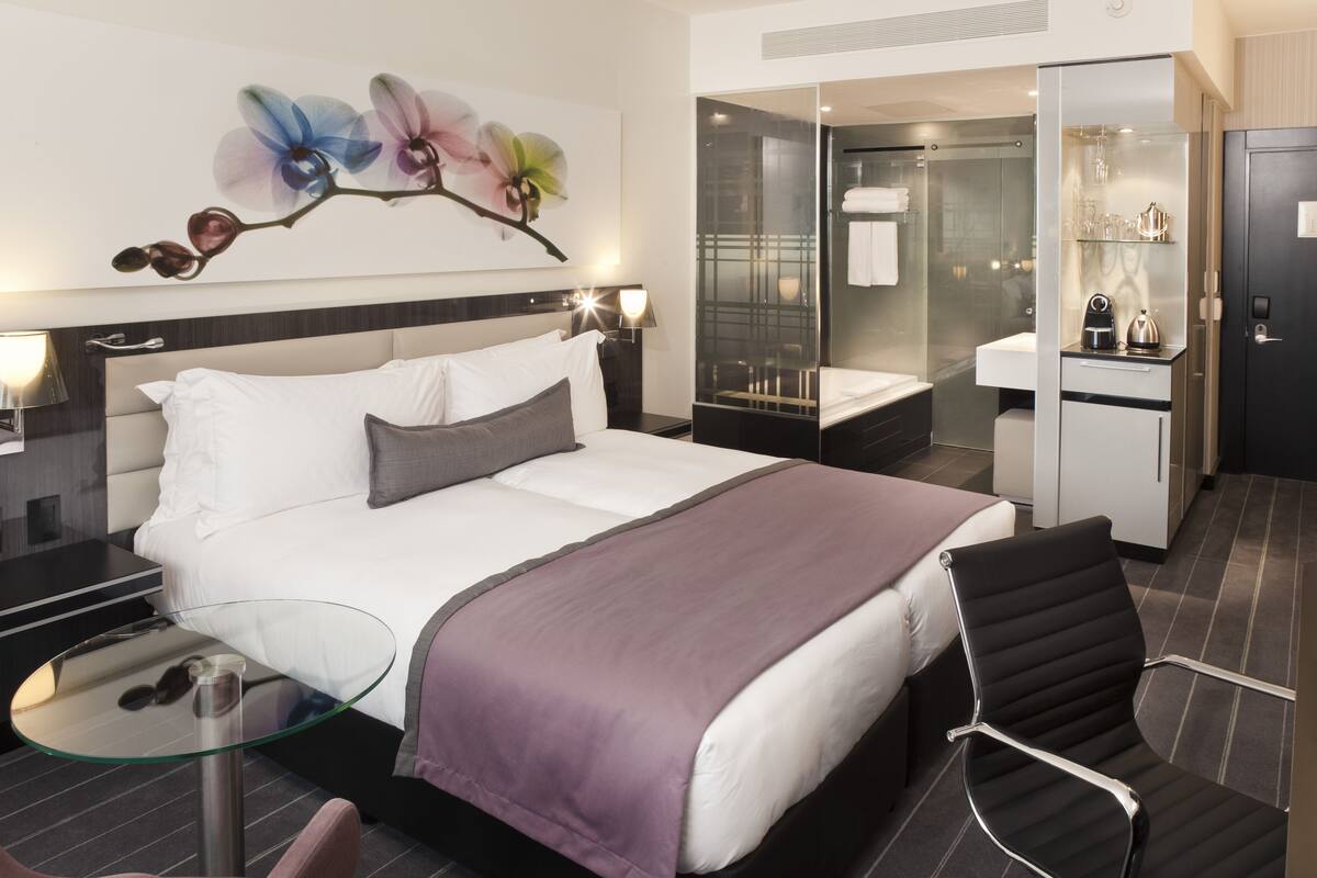 Une chambre d’hôtel moderne dotée d’un grand lit, d’une table en verre, d’une chaise, d’une salle de bain avec une douche en verre et d’un mini-bar.