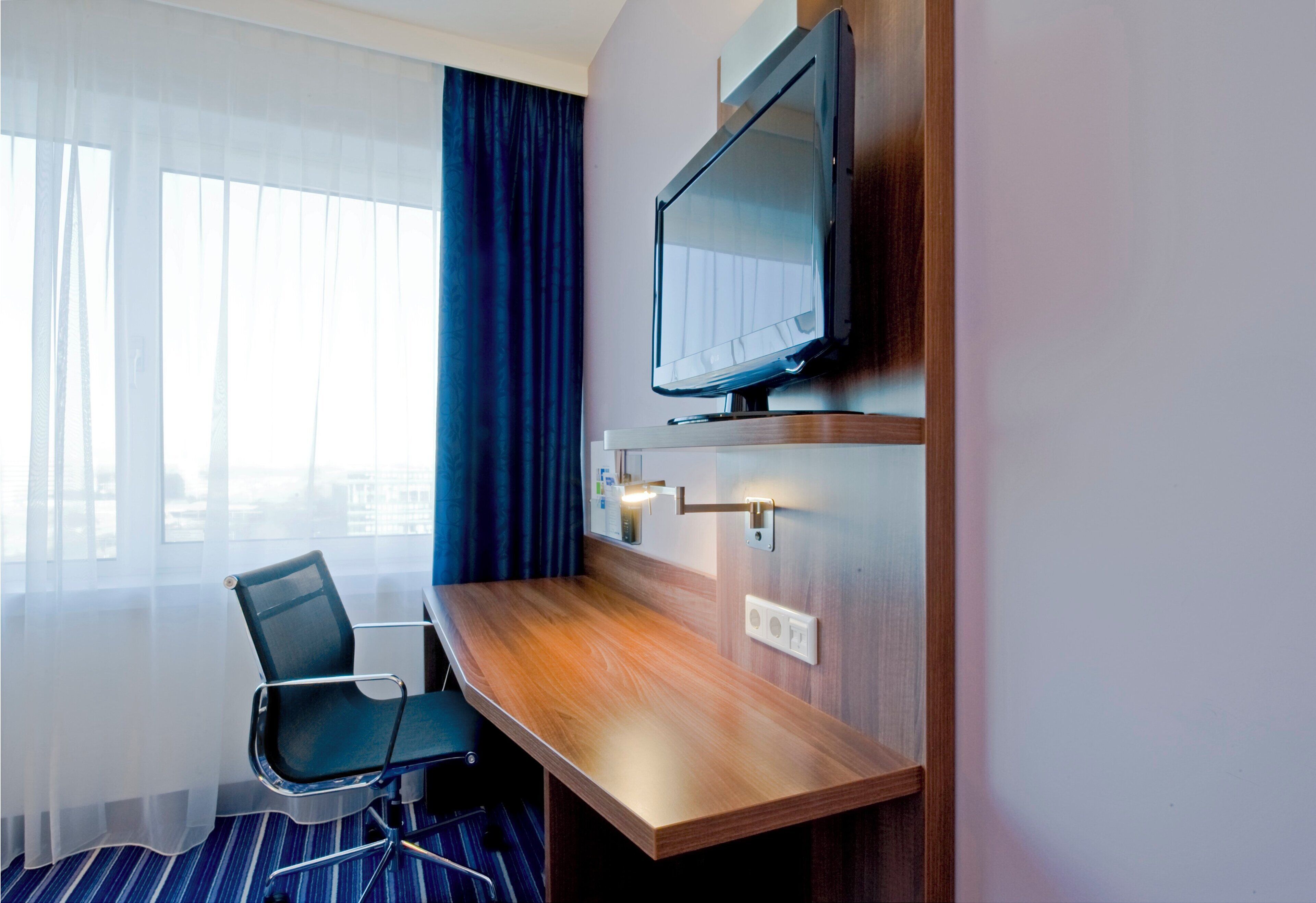 A hotel room with a wooden desk, a chair, a television mounted on the wall, and a window with sheer curtains.