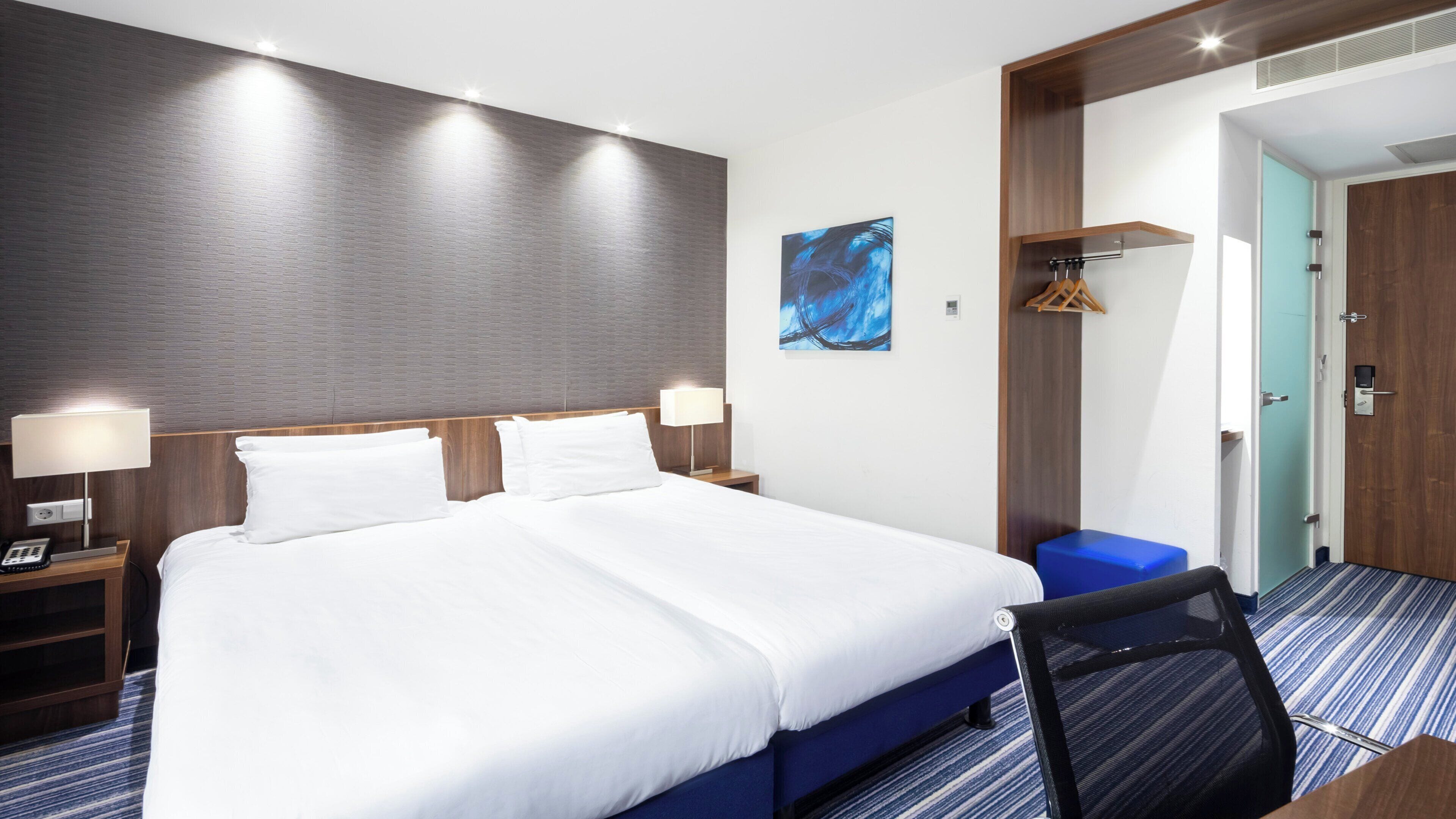 Ett hotellrum med en stor säng, sänglampor, en blå stol, en klädställning och en dörr till badrummet.
