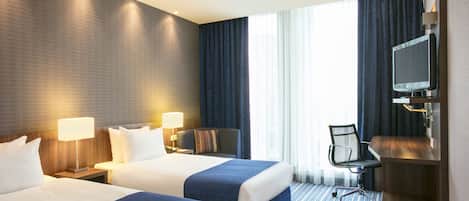 A hotel room with two beds, a desk, a chair, a television, and a large window with curtains.
