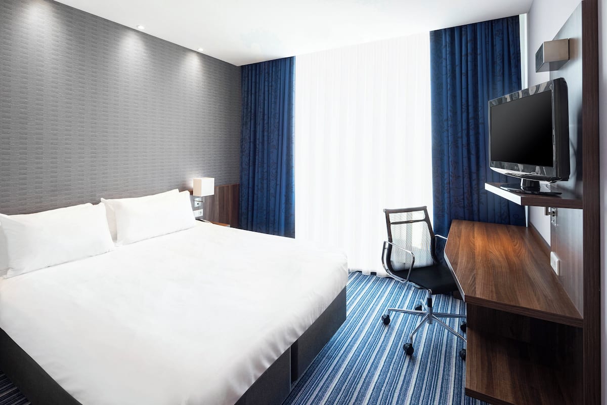A hotel room with a large bed, a desk with a flat-screen TV, a chair, and a desk lamp.