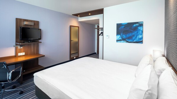 Habitación de hotel con cama, escritorio con televisor de pantalla plana, silla, un cuadro en la pared y una puerta que da a un baño.