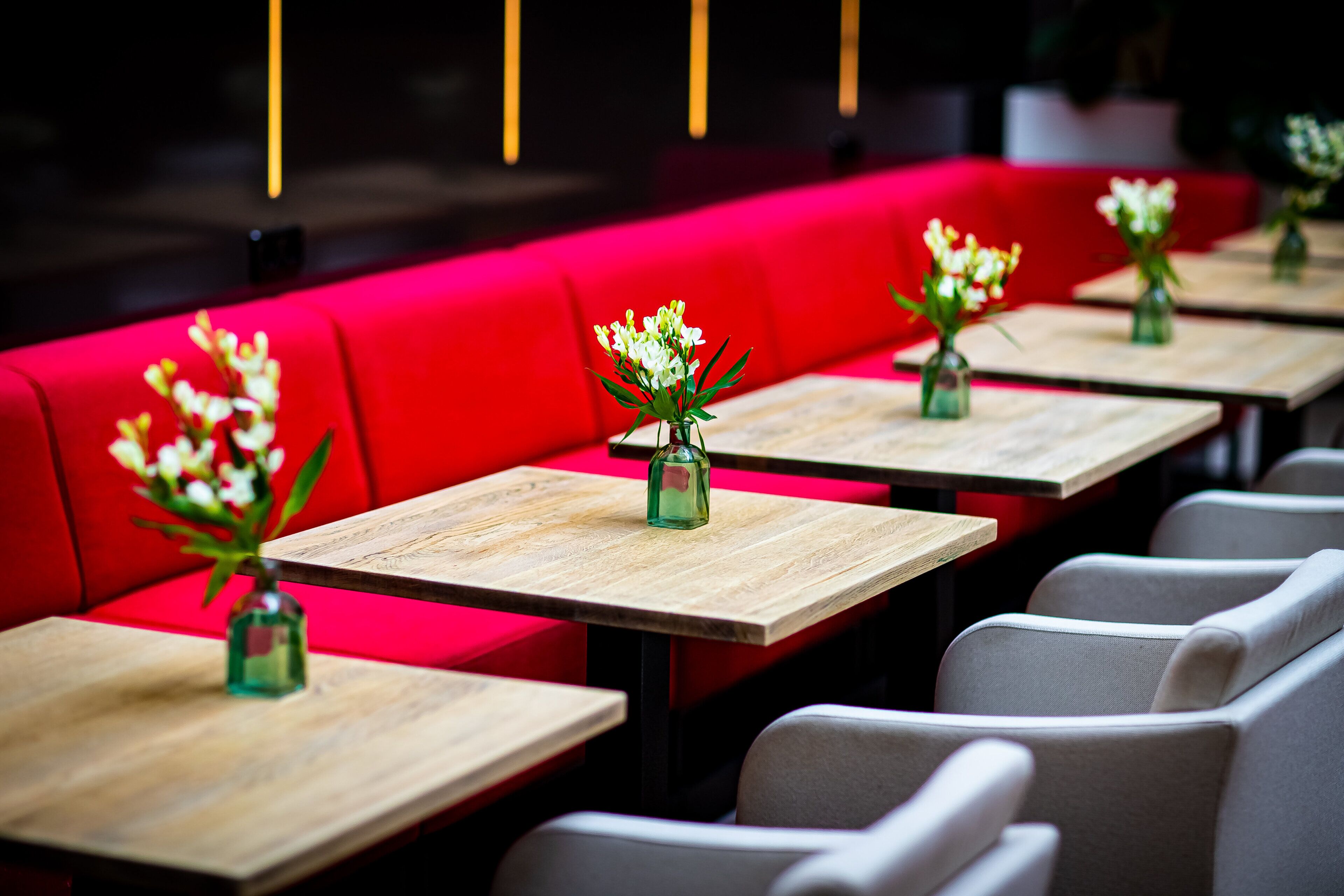 En rad bord med vaser fulla av blommor, röda sittplatser och grå stolar i en restaurangmiljö.