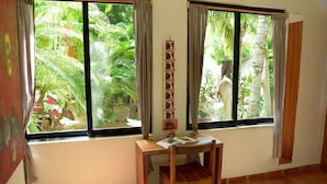 A room with a wooden desk, a chair, and a bed. There are two windows with curtains, and a view of greenery outside.