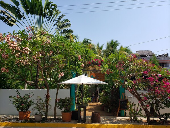 L’entrée d’un hôtel, avec un chemin menant à un espace de repos abrité, entouré d’une végétation tropicale luxuriante et d’une structure à toit de chaume.