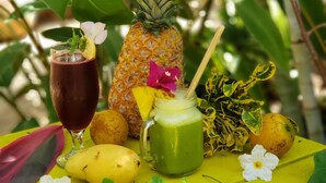 A tropical outdoor setting with a table featuring a pineapple, a glass of dark beverage, a green smoothie, and various fruits and flowers.