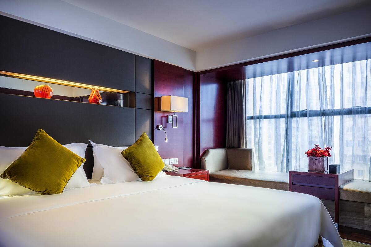 Ein Hotelzimmer mit einem großen Bett, einem Nachttisch, einem Sessel, einem Fenster mit Vorhängen und einer Wandregal mit Dekoration.