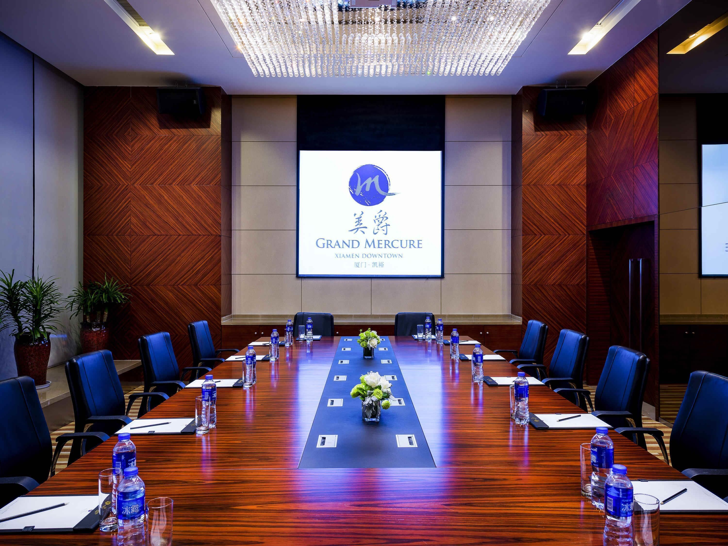 A conference room with a long table, chairs, water bottles, and a screen displaying 'GRAND MERCURY XIAMEN DOWNTOWN'.