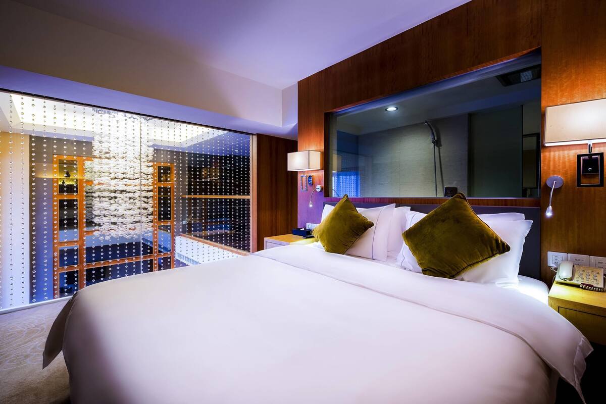 Ein Hotelzimmer mit einem großen Bett, einem Holz-Kopfteil, einem großen Spiegel und Blick auf eine Stadtlandschaft durch eine Glaswand.