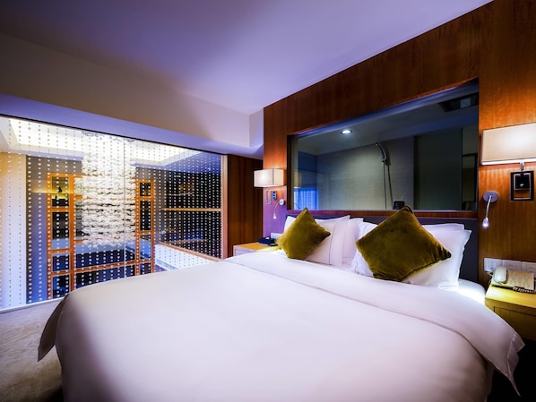 A hotel room with a large bed, a wooden headboard, a glass partition, and a view of the city.
