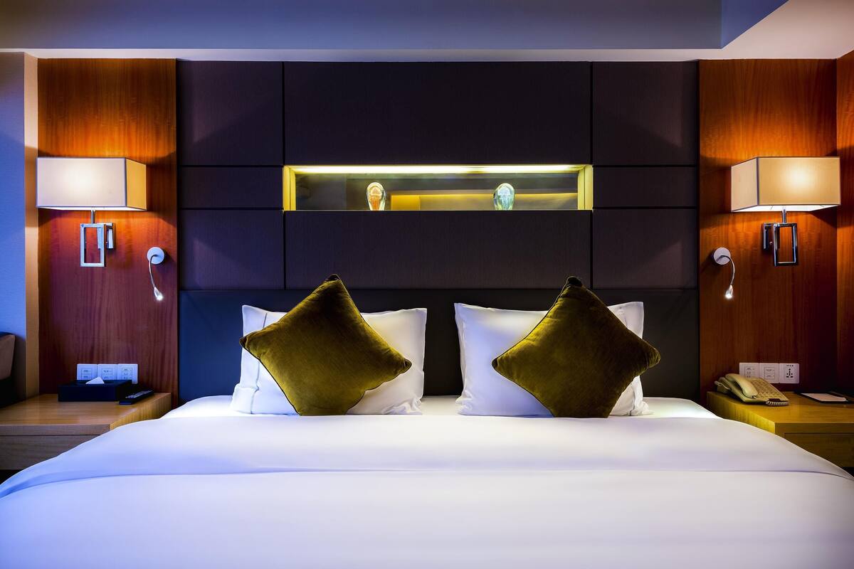 Ein Hotelzimmer mit einem großen Bett, zwei Nachttischlampen, einem Nachttisch, einem Telefon und einem an der Wand befestigten Spiegel.
