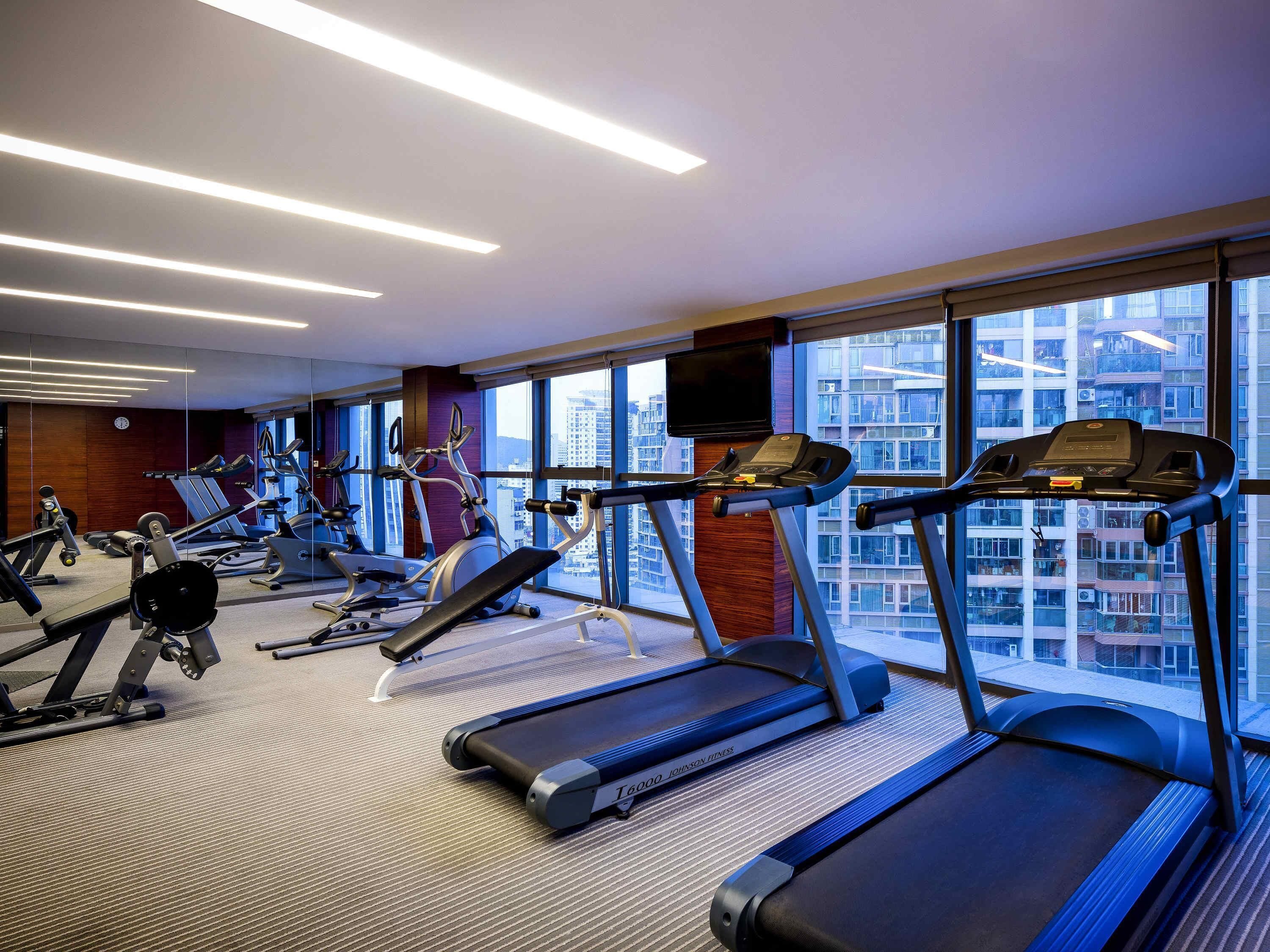 A modern gym with treadmills, elliptical machines, and weight equipment. Large windows offer a view of a cityscape.