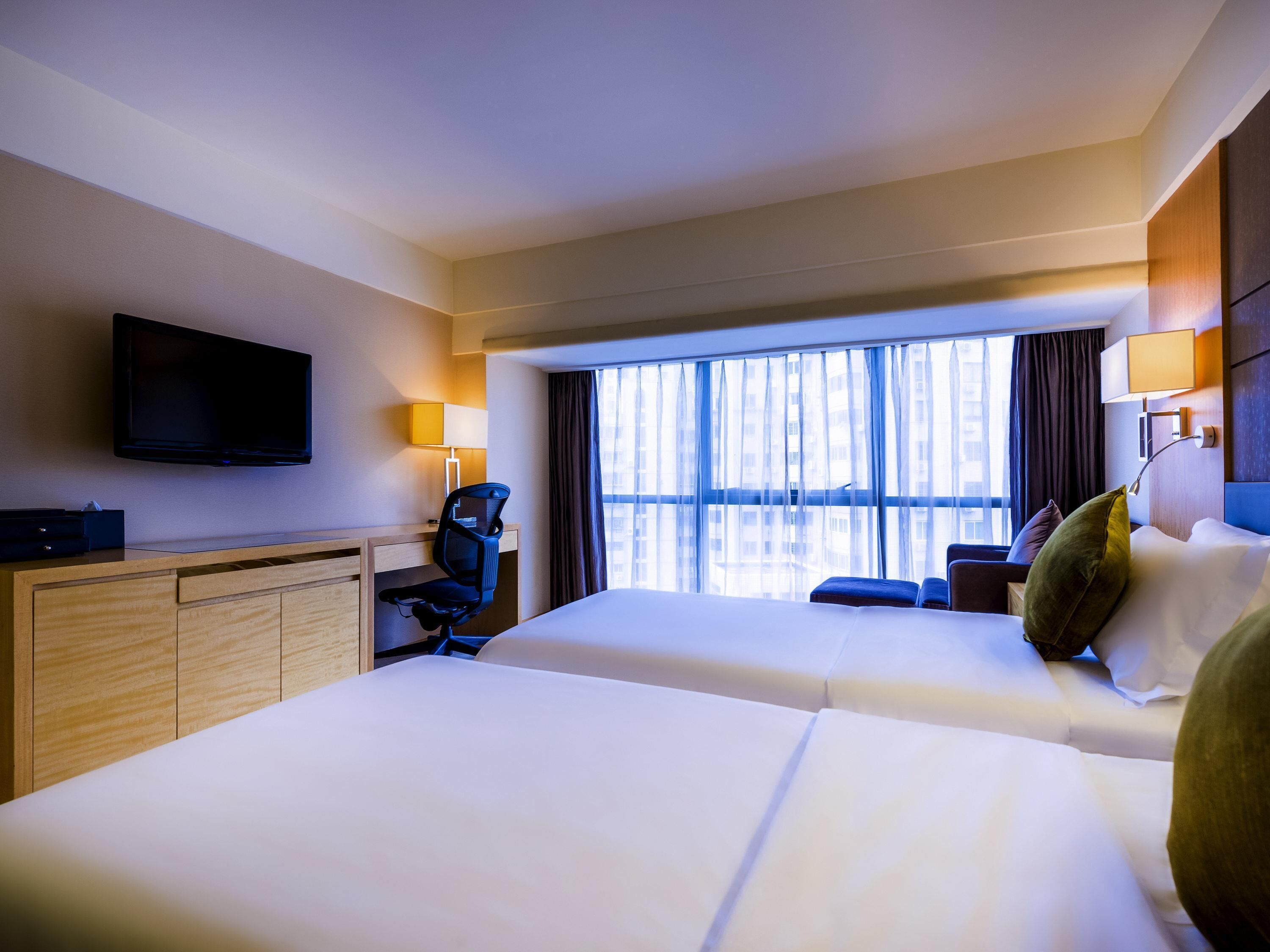 Quarto de hotel com duas camas, uma escrivaninha, uma cadeira, televisão e uma janela grande com cortinas.