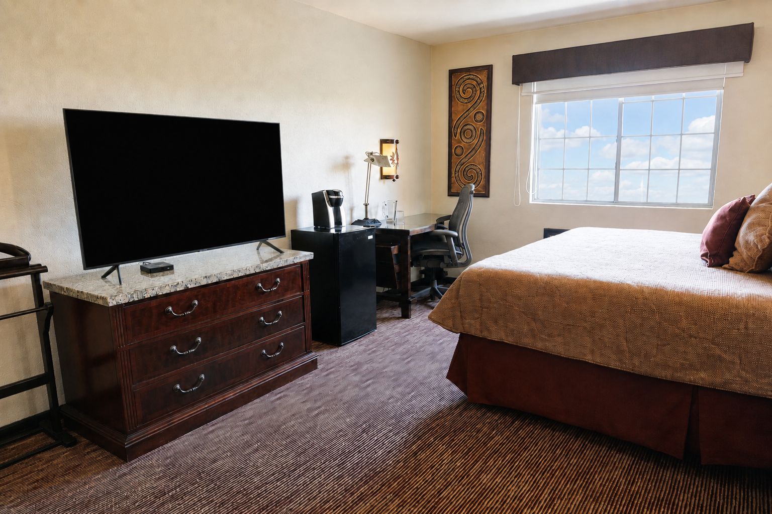 Habitación de hotel con un televisor de pantalla plana grande, un aparador de madera, un escritorio con silla, una cama con colcha marrón y una ventana con vista al cielo.
