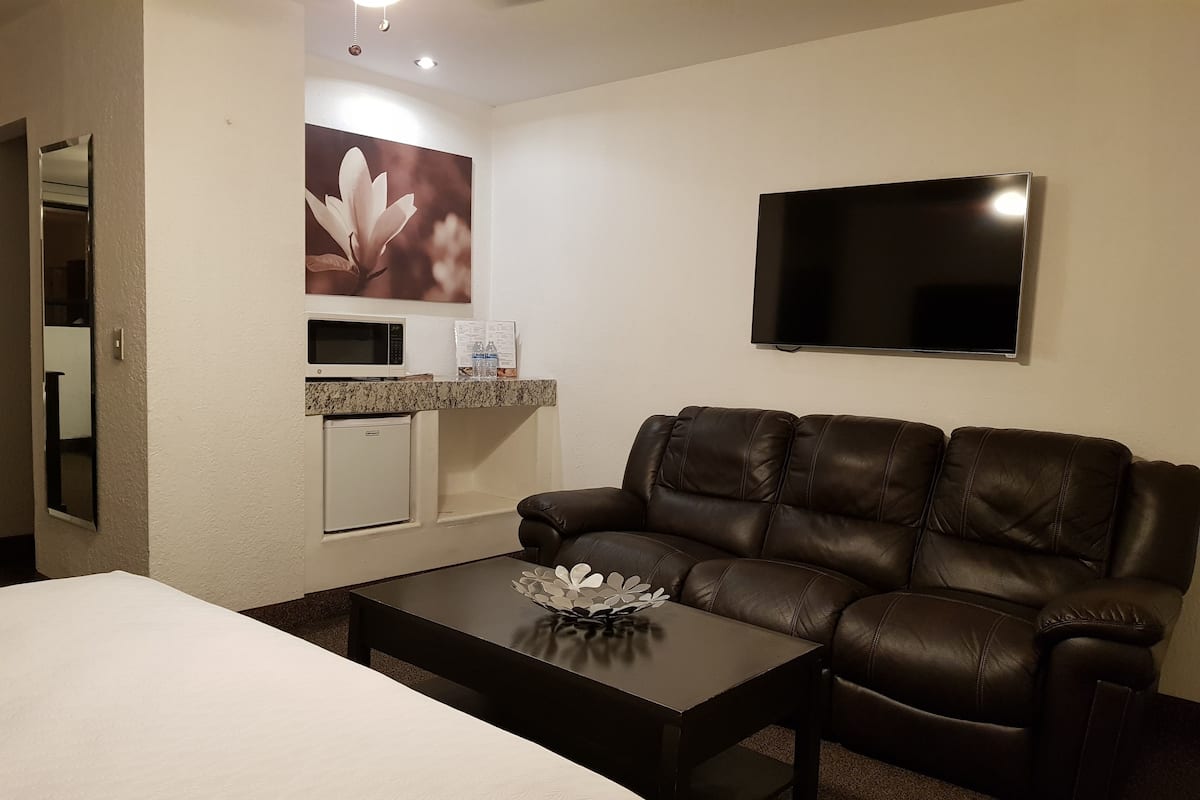 A hotel room with a bed, a leather sofa, a small kitchenette, a flat-screen TV, and a decorative flower vase.