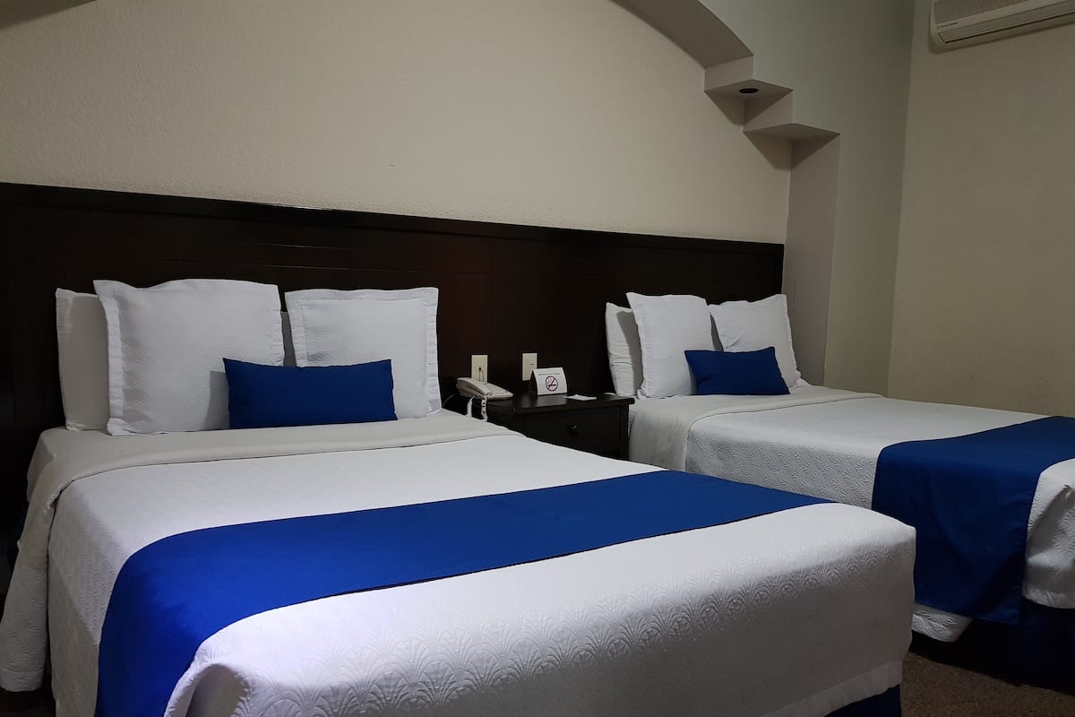 Two single beds with white and blue bedding, a dark headboard, a nightstand with a phone, and a wall-mounted air vent.