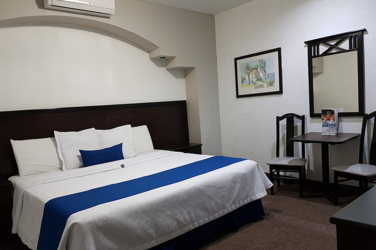 A neatly made bed with a blue and white bedspread, a wooden headboard, and a small table with a chair beside it.