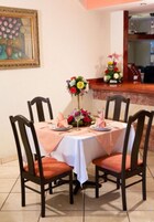 Un comedor con una mesa redonda dispuesta para cuatro personas, rodeada de sillas de madera con asientos acolchados. Un mantel blanco cubre la mesa, y hay centros de mesa florales sobre ella.