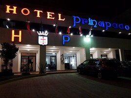 Entrada de un hotel con letreros iluminados, un emblema de corona y banderas.